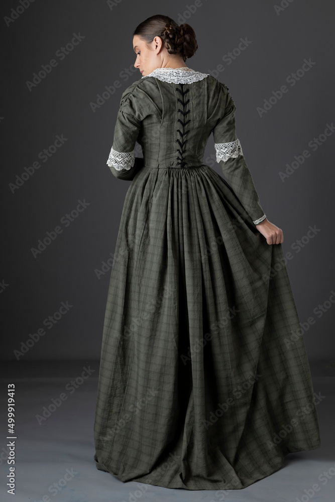 A Victorian working class woman wearing a checked bodice and skirt and standing alone against a studio backdrop