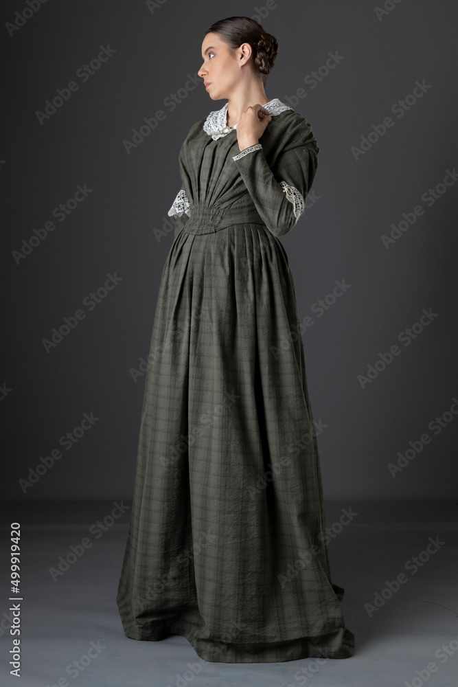 A Victorian working class woman wearing a checked bodice and skirt and standing alone against a studio backdrop A Victorian working class woman wearing a checked bodice and skirt and standing alone against a studio backdrop