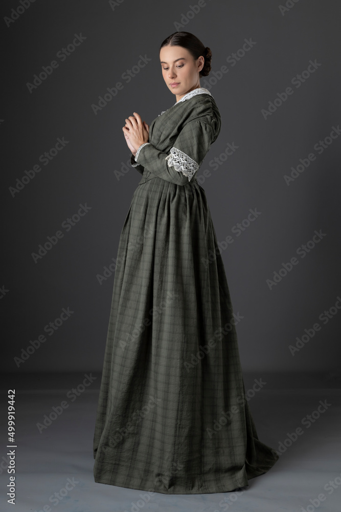 A Victorian working class woman wearing a checked bodice and skirt and standing alone against a studio backdrop