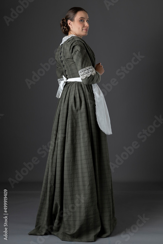 A Victorian woman wearing a dark green checked bodice and skirt and standing alone against a studio backdrop