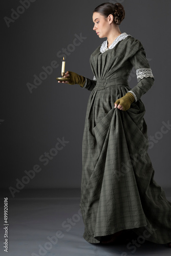 A Victorian working class woman wearing a dark green checked bodice and skirt and carrying a candle in a darkened room