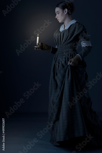 A Victorian working class woman wearing a dark green checked bodice and skirt and carrying a candle in a darkened room
