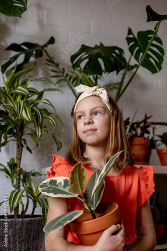 Teenage girl in coral dress sits in room and holds ficus in her hands.