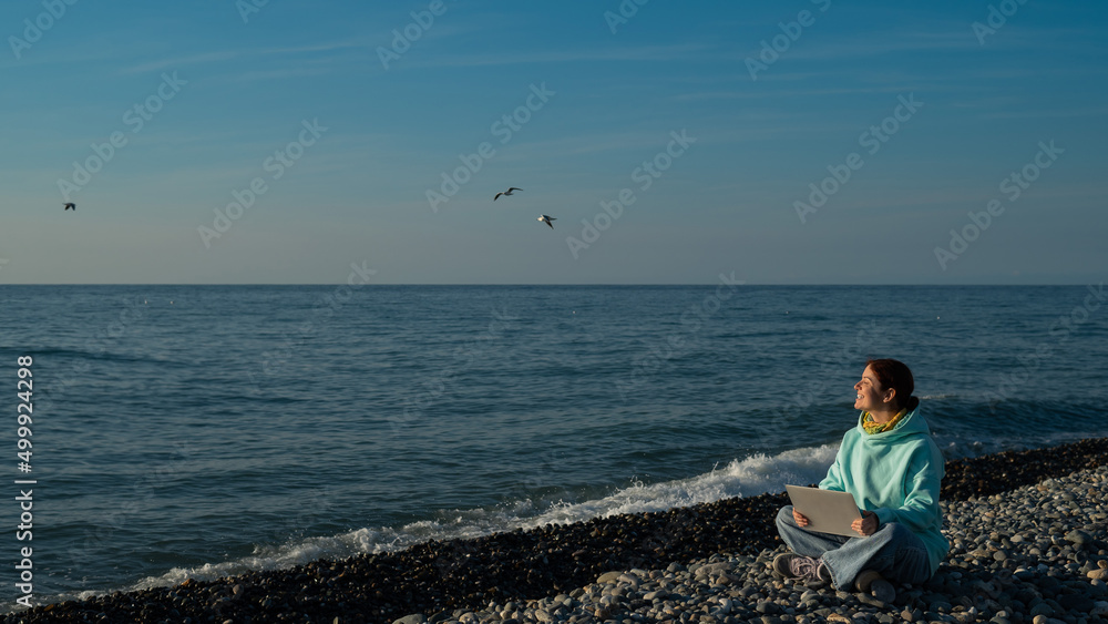 Happy caucasian woman working on a laptop while sitting on a pebble beach.