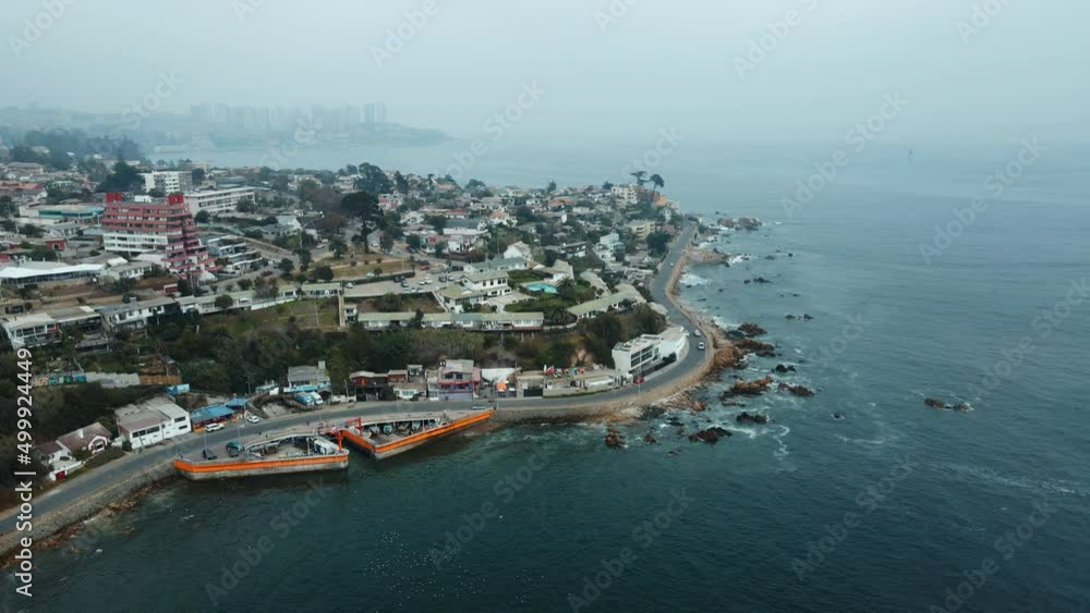 Aerial orbit of coastal road between Concon hillside city buildings and sea waves hitting rocky shore on an overcast day, La Boca beach, Chile Aerial orbit of coastal road between Concon hillside city buildings and sea waves hitting rocky shore on an overcast day, La Boca beach, Chile