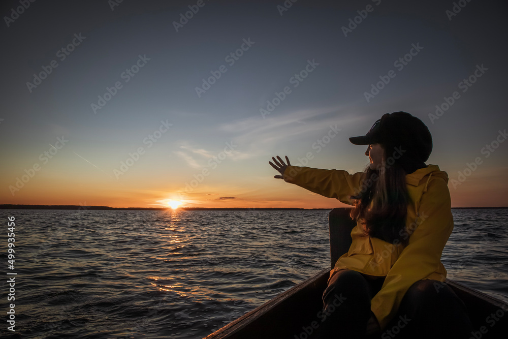 Young woman is waving goodbye to the sunset while sitting in wooden boat