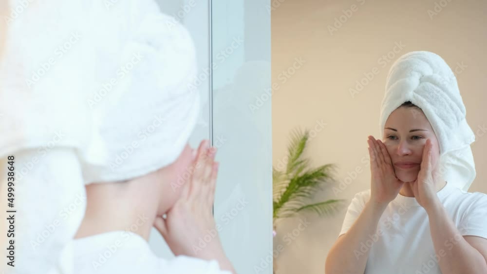 Young woman with a white towel on her head doing self-massage of the face near mirror. Young woman with a white towel on her head doing self-massage of the face near mirror.
