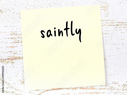 Yellow sticky note on wooden wall with handwritten word saintly