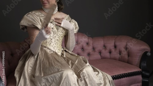 A Victorian woman wearing a gold ball gown and sitting on a pink sofa fanning herself as if in a ball room/ A candle light filter has been added photo
