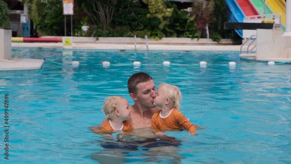 In a large pool with clear blue water, the father keeps two twin girls, kisses the girls in turn and the three of them enjoy swimming and smile sweetly. Happy holidays with kids.