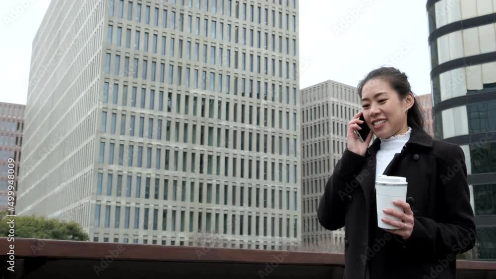 Low angle tracking shot of glad Chinese female entrepreneur with zero waste cup of coffee to go. She is smiling and speaking on cellphone while strolling on city street in morning Low angle tracking shot of glad Chinese female entrepreneur with zero waste cup of coffee to go. She is smiling and speaking on cellphone while strolling on city street in morning