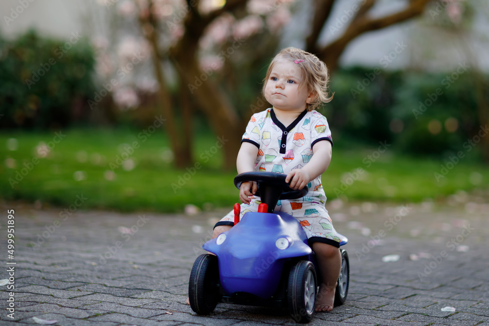 Cute little baby girl playing with blue small toy car in garden of home or nursery. Adorable beautiful toddler child with blossoming magnolia on background Cute little baby girl playing with blue small toy car in garden of home or nursery. Adorable beautiful toddler child with blossoming magnolia on background