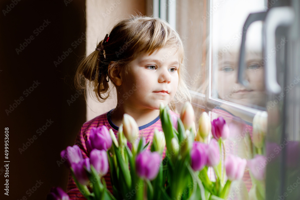 Naklejka premium Little girl sitting by window with tulip flowers bouquet. Happy child, indoors. Mother's day, valentine's day or birthday concept.