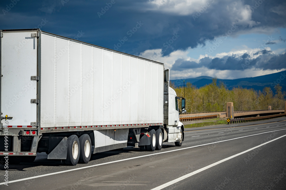 Bright white big rig semi truck transporting cargo in dry van semi trailer driving on the highway road with dark stormy sky