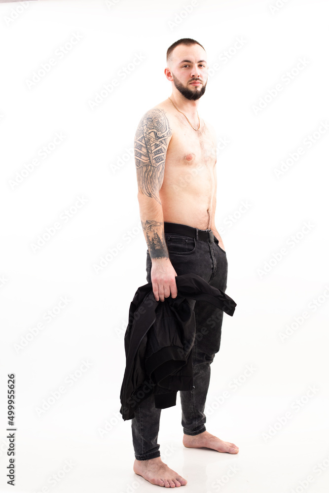 Portrait of young gorgeous bearded man with short dark hair wearing black jeans, golden chain with bare torso standing. Portrait of young gorgeous bearded man with short dark hair wearing black jeans, golden chain with bare torso standing.