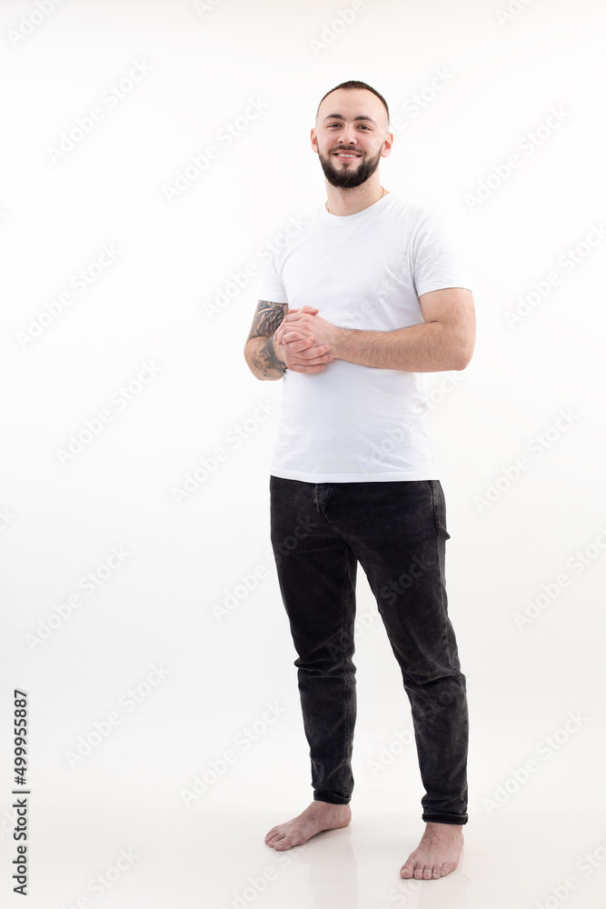 Portrait of young happy smiling bearded man with short dark hair in white T-shirt, black jeans, stand joining hands. Portrait of young happy smiling bearded man with short dark hair in white T-shirt, black jeans, stand joining hands.