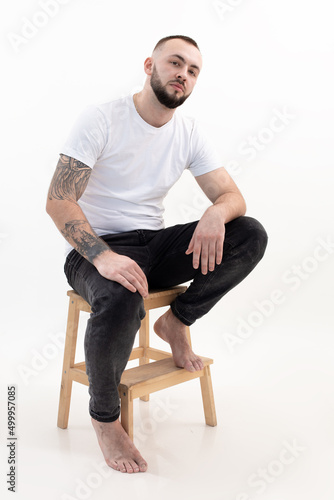 Young handsome bearded man with short dark hair in white T-shirt, black jeans, sit on step-ladder putting foot on stair.