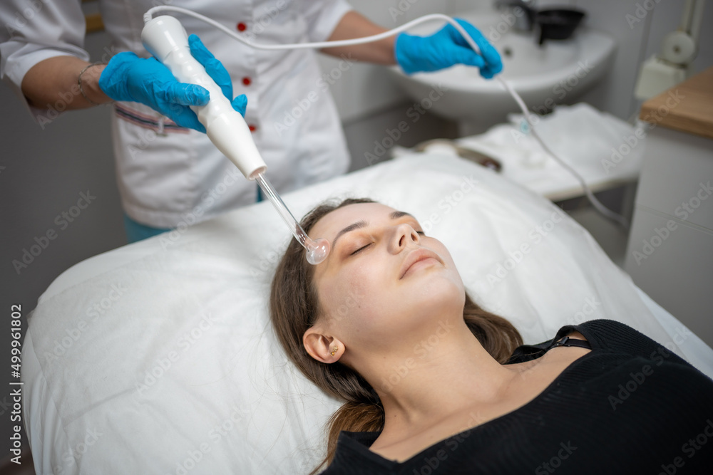 Fototapeta premium Beautician uses the darsonval device on the client's face. Professional skin care in the salon. The concept of cosmetology and rejuvenation.