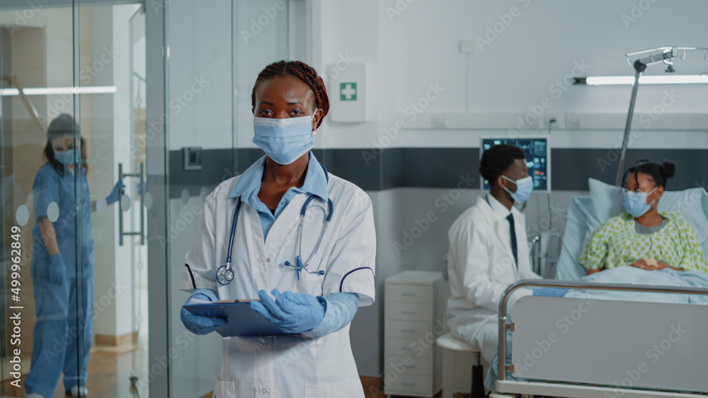 Fototapeta premium Portrait of woman working as doctor with white coat and stethoscope, standing in hospital ward. Medical specialist wearing face mask being ready to treat ill patient with medicine