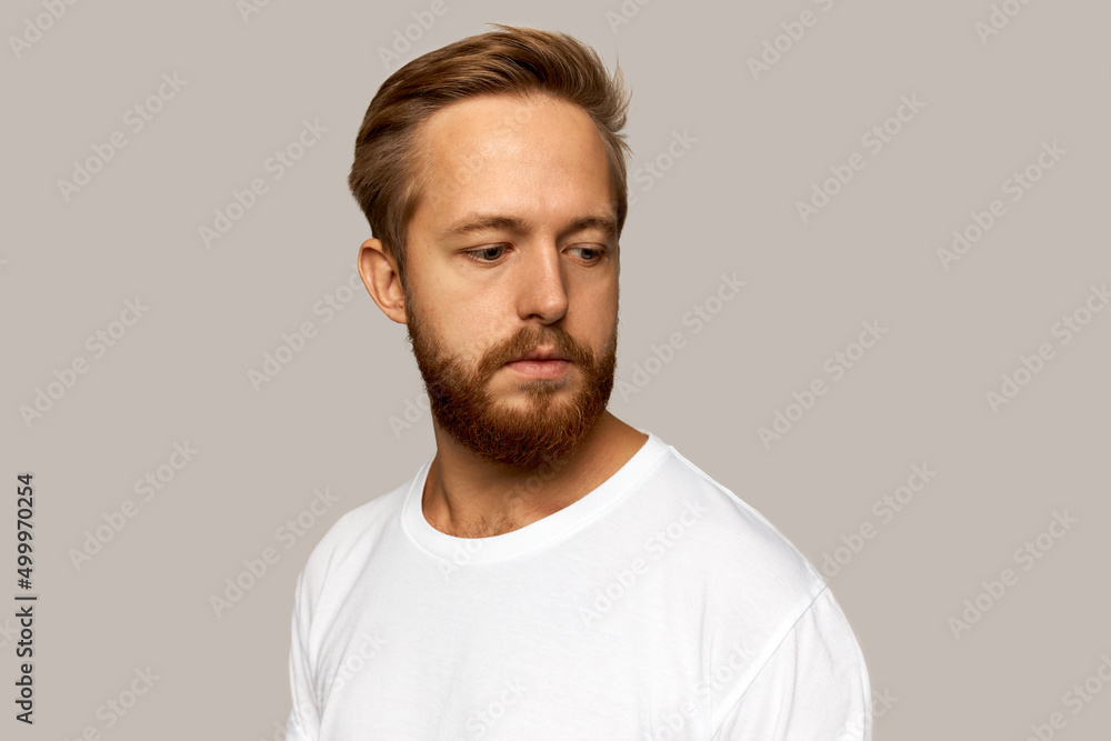 Picture of thoughtful pensive white-skinned bearded male in casual white t-shirt standing against studio wall looking down, trying to remember something important, having stylish haircut