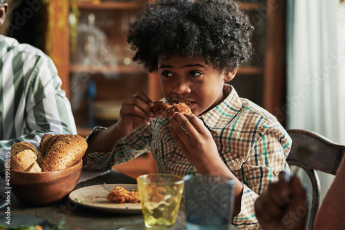 African little boy sitting at dining table and eating roast chicken during dinner with his family