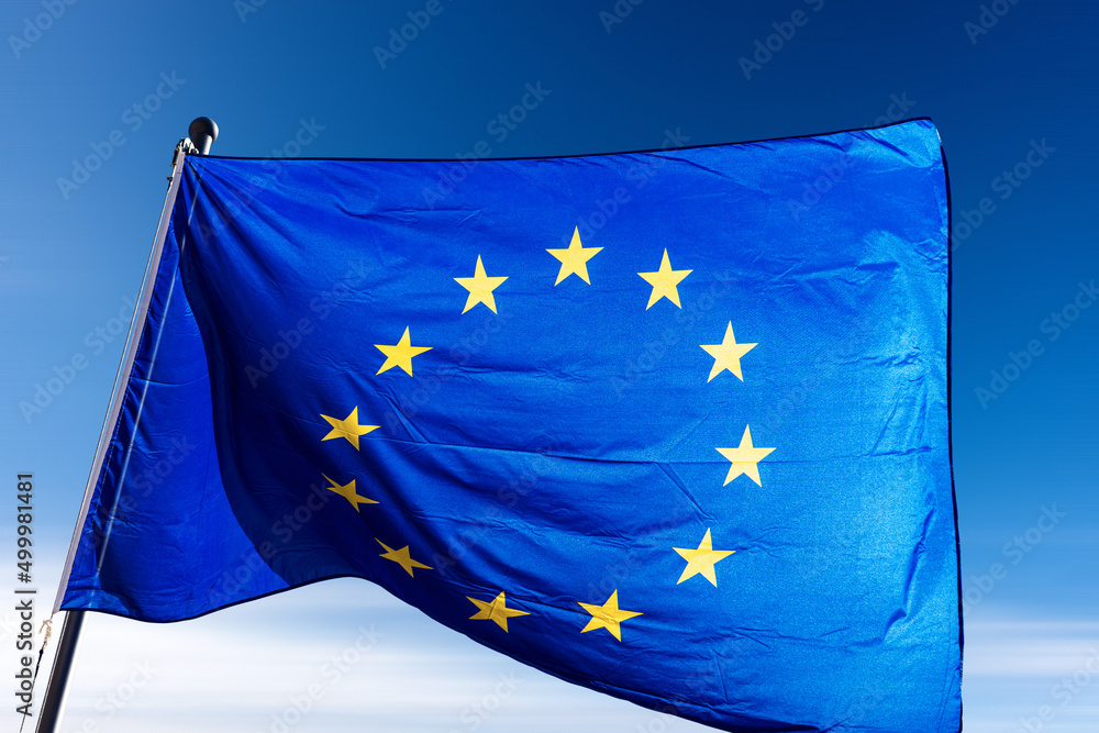 Close-up of an European Union flag waving against a clear blue sky with clouds and copy space. Close-up of an European Union flag waving against a clear blue sky with clouds and copy space.