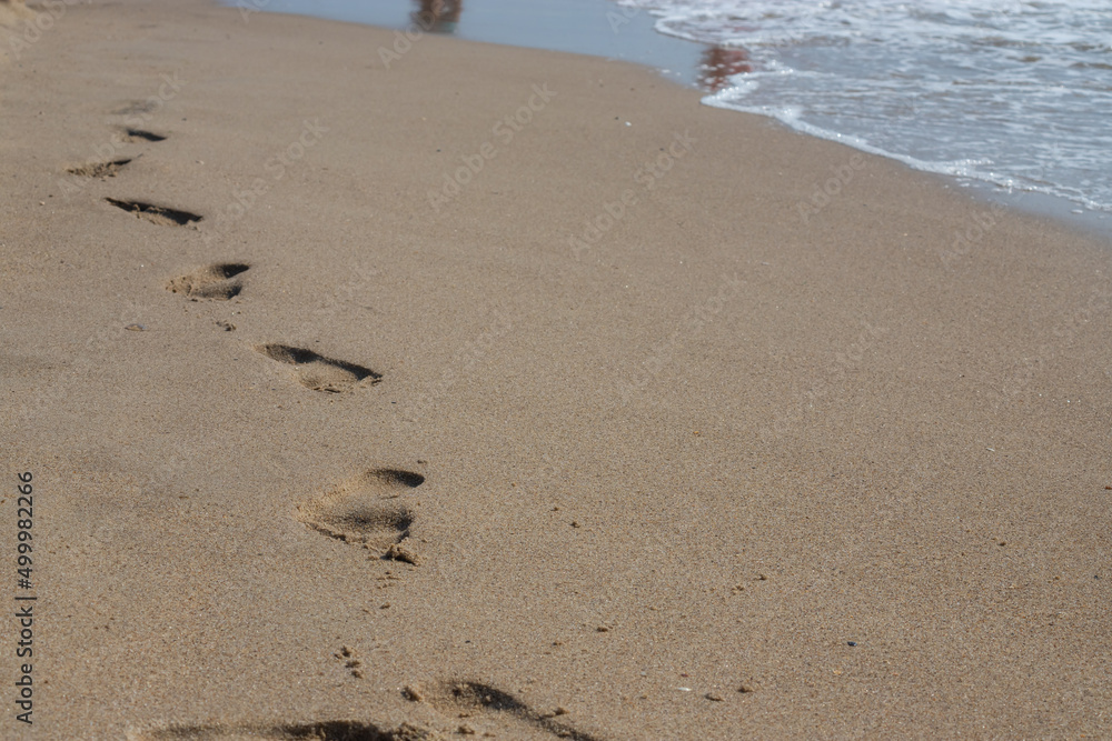 Footprints in the sand beach. Footprints in the sand against a sea wave. Footprints on a sunny day with golden sand, beach, wave and footsteps Footprints in the sand beach. Footprints in the sand against a sea wave. Footprints on a sunny day with golden sand, beach, wave and footsteps