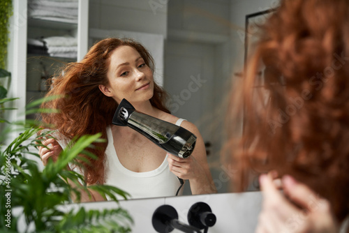 Obraz na plátne Redhead caucasian woman drying hair in the bathroom in front of mirror