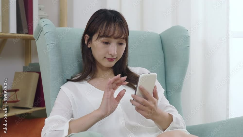 carefree japanese female is tapping and sliding on the phone screen while enjoying social media feeds on the sofa in the apartment. carefree japanese female is tapping and sliding on the phone screen while enjoying social media feeds on the sofa in the apartment.