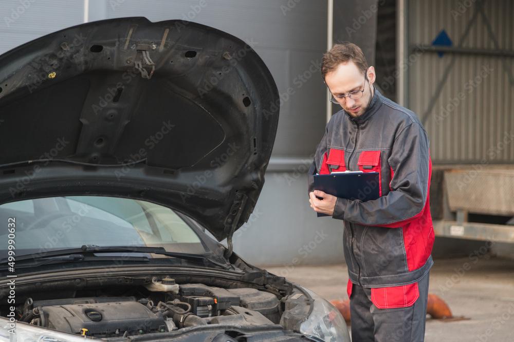 Caucasian mechanic in uniform making notes during car maintenance at car repair station Caucasian mechanic in uniform making notes during car maintenance at car repair station