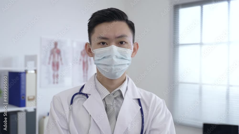 shoulder shot asian male doctor is adjusting his face mask and looking at the camera with a serious expression at a clinic office. shoulder shot asian male doctor is adjusting his face mask and looking at the camera with a serious expression at a clinic office.