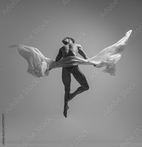 Fototapeta Naklejka Na Ścianę i Meble -  Flight. Black and white portrait of graceful muscled male ballet dancer dancing with fabric, cloth isolated on grey studio background. Grace, art, beauty concept. Weightless, flexible.