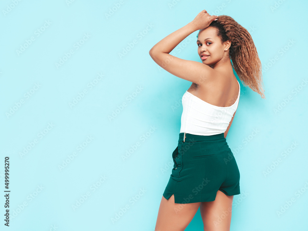Beautiful black woman with afro curls hairstyle. Smiling model dressed in summer shorts. Sexy carefree female posing near blue wall in studio. Tanned and cheerful Beautiful black woman with afro curls hairstyle. Smiling model dressed in summer shorts. Sexy carefree female posing near blue wall in studio. Tanned and cheerful