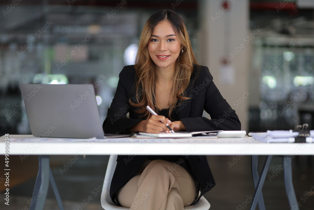 Portrait of young asian businesswoman smiling and looking at camera, thinking and writing on notebook while sitting at office desk in modern office. Portrait of young asian businesswoman smiling and looking at camera, thinking and writing on notebook while sitting at office desk in modern office.
