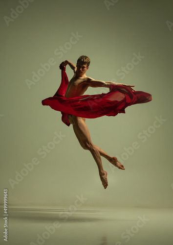 One young muscled man, flexible ballet dancer in action with red fabric, cloth isolated on olive color background. Theater, emotions, grace, art, beauty concept.