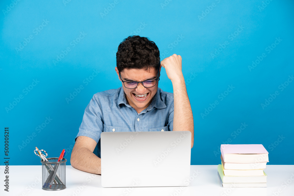 Happy hispanic teenager celebrating good news or results in front of laptop isolated on blue background. Happy hispanic teenager celebrating good news or results in front of laptop isolated on blue background.