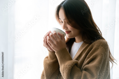 Young asian woman standing beside window and holding mug in bedroom at home, She drinking milk after wake up in the morning photo