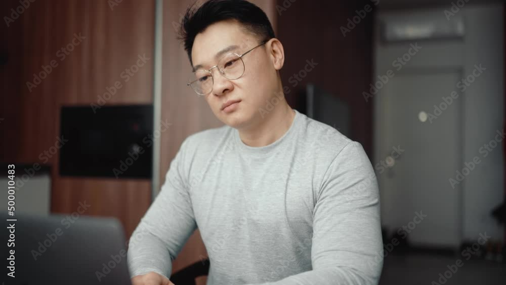 Asian serious man wearing eyeglasses working on laptop at home Asian serious man wearing eyeglasses working on laptop at home