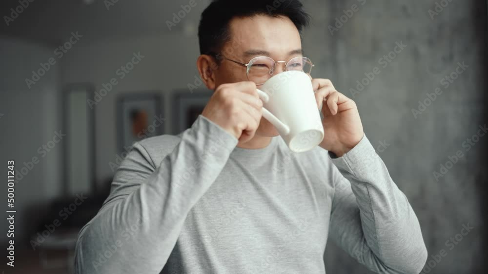 Asian smiling man wearing eyeglasses talking on mobile and drinking coffee at home Asian smiling man wearing eyeglasses talking on mobile and drinking coffee at home