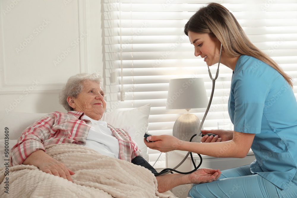 Young caregiver measuring blood pressure of senior woman in room. Home health care service Young caregiver measuring blood pressure of senior woman in room. Home health care service