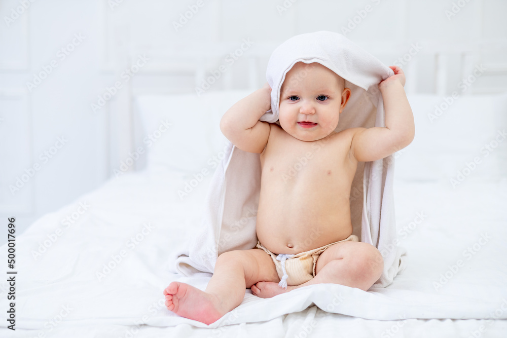 a baby in a white towel after bathing in a bathtub on a bright bed at home smiles, the concept of hygiene and washing a baby in a white towel after bathing in a bathtub on a bright bed at home smiles, the concept of hygiene and washing