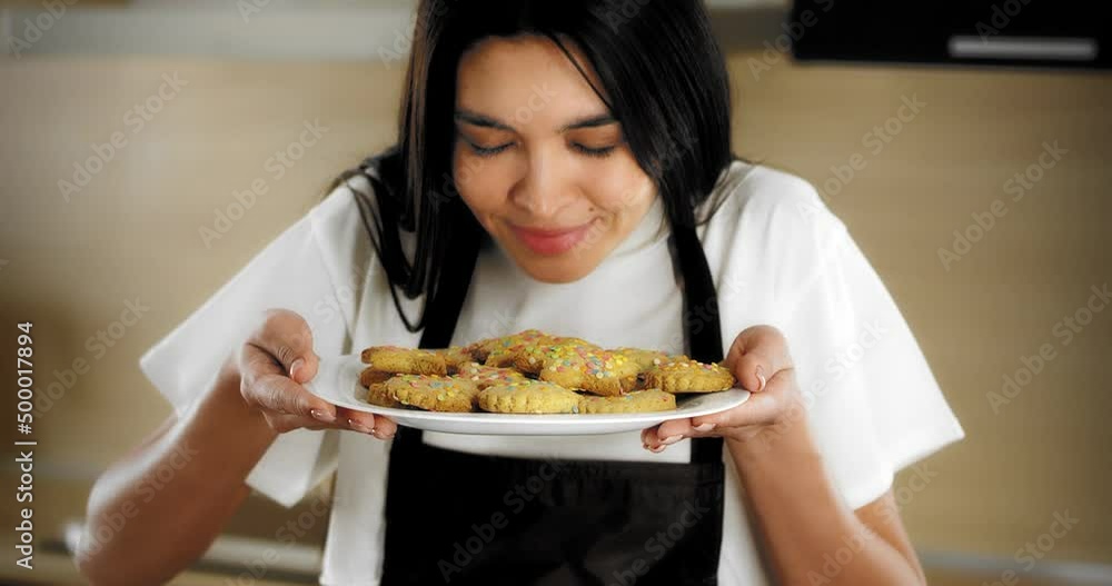 Pretty woman mixed-race brunette dressed in a white T-shirt and a brown apron sprinkles decorations on shortbread cookies and gingerbread lying on parchment on a baking sheet Pretty woman mixed-race brunette dressed in a white T-shirt and a brown apron sprinkles decorations on shortbread cookies and gingerbread lying on parchment on a baking sheet