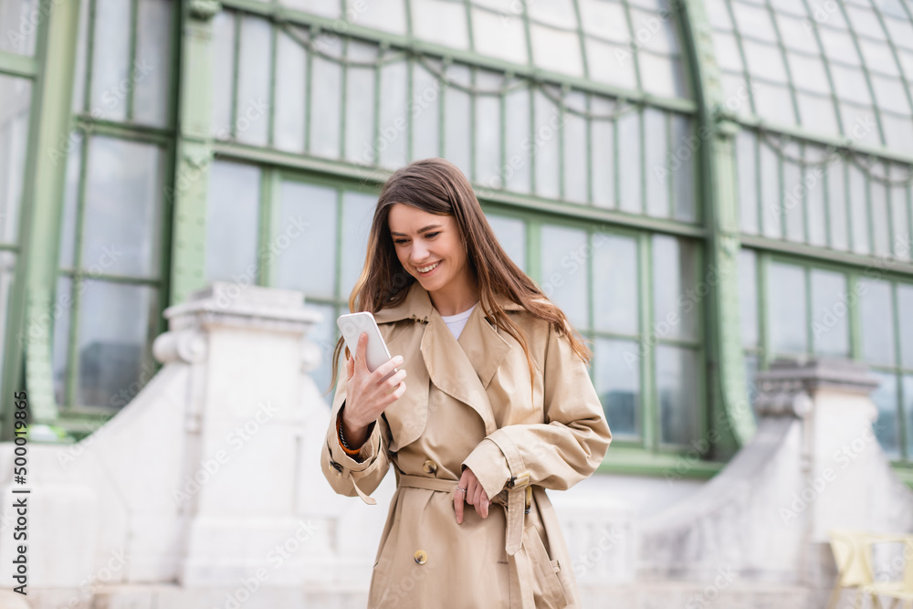 happy young woman in trench coat looking at smartphone near european building.