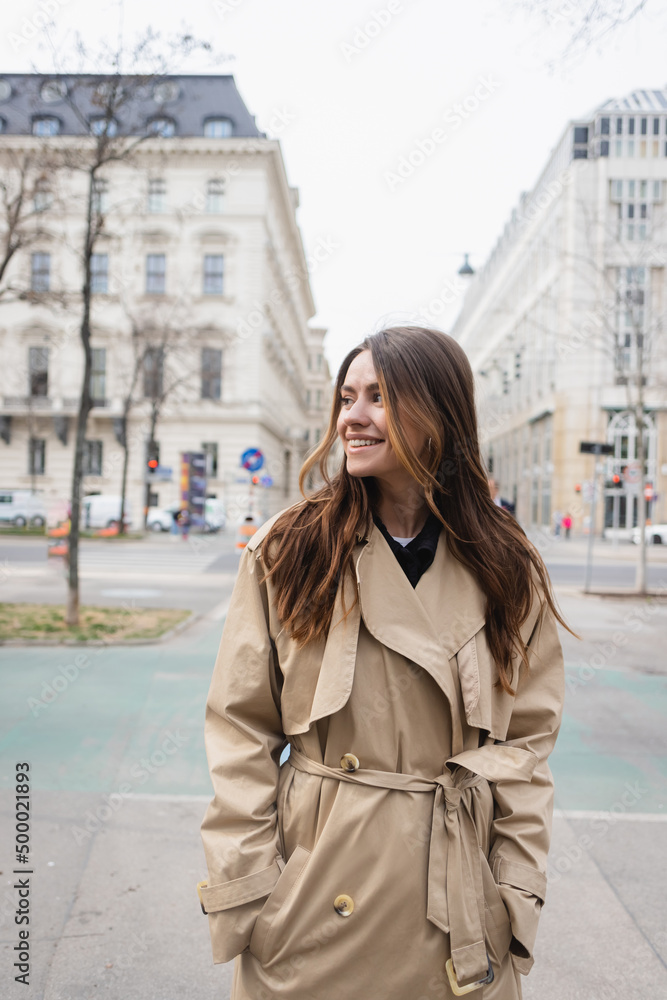 cheerful young woman in stylish trench coat standing with hands in pockets on street of european city. cheerful young woman in stylish trench coat standing with hands in pockets on street of european city.