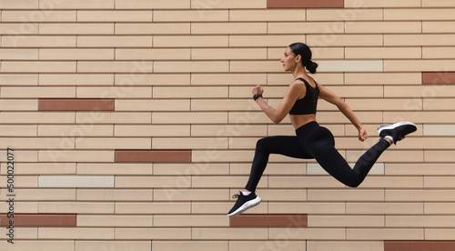 Serious young arabic female athlete in sportswear runs, frozen in air on brick wall background, profile, panorama