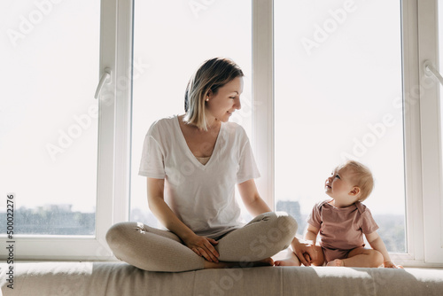 Fotografi Mother and little daughter sitting at home on a windowsill by a big window, looking at each other, chatting and smiling