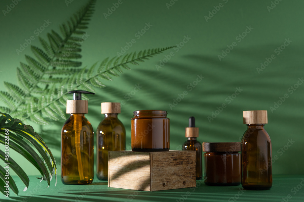 Bottles for cosmetics and cream on a pedestal, green background with leaves and shadow. Brown glass jars with wooden lids. Eco concept. Container for shampoo, oil. Bottle with dispenser and pipette. Bottles for cosmetics and cream on a pedestal, green background with leaves and shadow. Brown glass jars with wooden lids. Eco concept. Container for shampoo, oil. Bottle with dispenser and pipette.