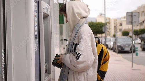 Girl in hoodie goes to an ATM on the street to withdraw cash. Banking service and cash withdrawal concept