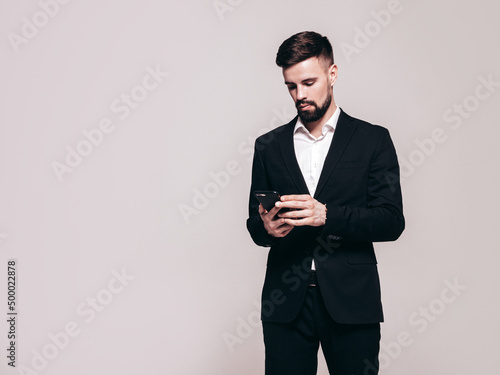 Handsome confident stylish hipster lambersexual model. Sexy modern man dressed in elegant black suit. Fashion male posing in studio. Holding smartphone. Looking at cellphone screen. Using apps