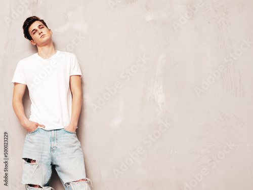 Portrait of handsome confident stylish hipster lambersexual model.Man dressed in white t-shirt and jeans. Fashion male posing in studio near grey wall